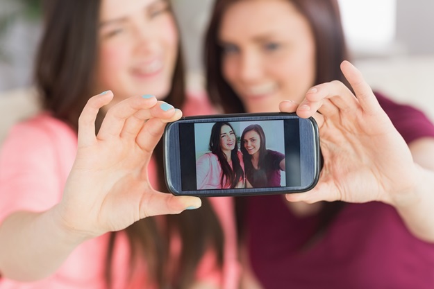 Two happy teenage girls sitting on a sofa taking a photo of themselves with a mobile phone in a living room