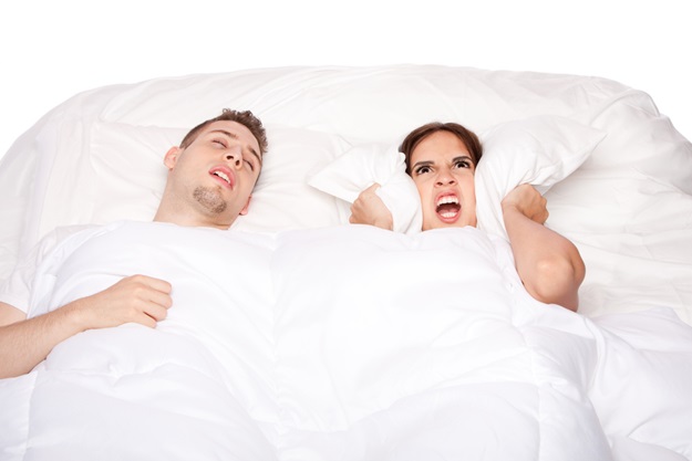 Couple laying asleep in bed while man snores and woman holds pillow over ears.